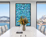 Bright contemporary interior featuring Listen Learn Teach, a large framed Aboriginal artwork in blue, turquoise, black, and white dot patterns displayed above a light wood cabinet beside tall sunlit windows